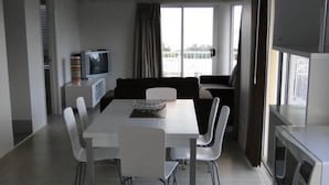 A modern, minimalist living room with a dining area. It features a flat-screen TV, a sofa, and a dining table with chairs.