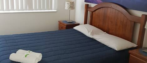 A neatly made bed with a blue quilt, a white pillow, and two rolled towels on the bed. There is a wooden nightstand with a lamp beside the bed. A ceiling fan is visible above the bed, and a large painting of a sunset is on the wall.