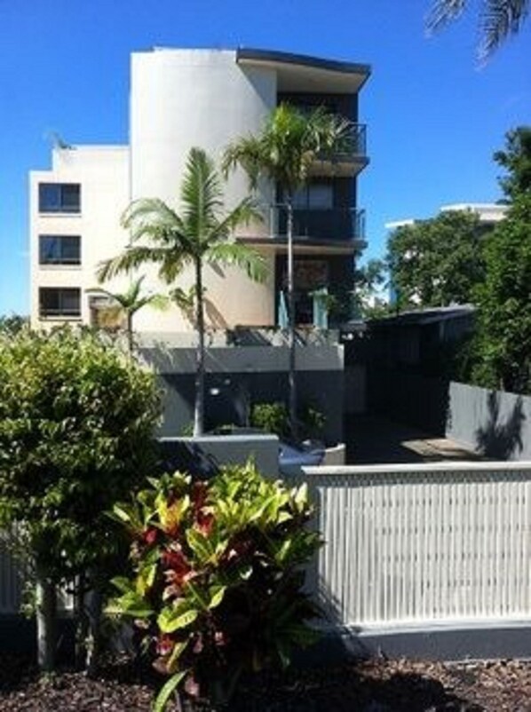 Il s’agit d’un bâtiment moderne doté d’un toit plat, de nombreuses fenêtres et d’un balcon. On peut voir divers arbustes et plantes au premier plan.