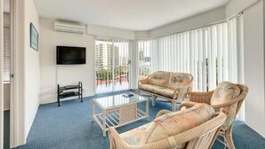 A living room with a flat-screen TV, wicker furniture, a glass coffee table, and large windows with blinds.