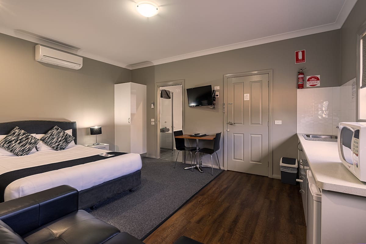 A compact hotel room with a bed, a desk, a chair, a microwave, and a small kitchenette.