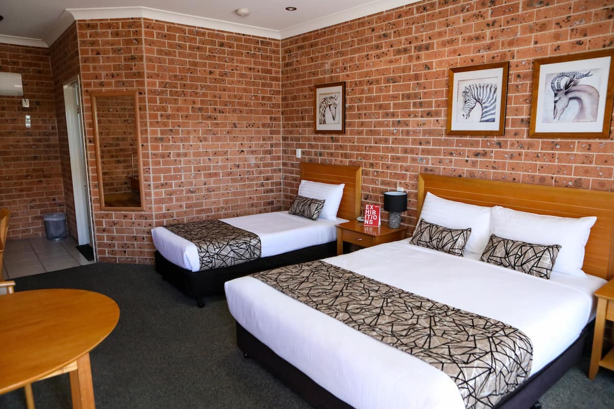 A hotel room with two beds, a wooden headboard, a small table, and a brick wall with framed artwork.