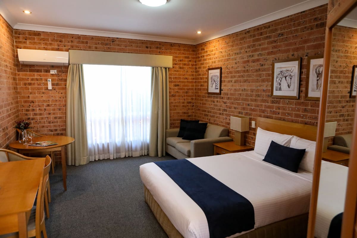 A hotel room with a brick wall, a bed with a blue bedspread, a wooden desk, a chair, a sofa, and a window with curtains.