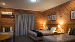 A hotel room with two beds, a desk, and a chair. There is a brick wall, a window with curtains, and a wall-mounted air conditioner. - Akuna Motor Inn and Apartments (Dubbo)