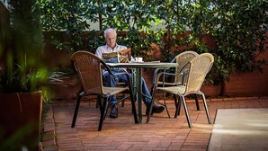 A man sitting at a table with a magazine, surrounded by wicker chairs in an outdoor setting with brick flooring and greenery. - Akuna Motor Inn and Apartments (Dubbo)