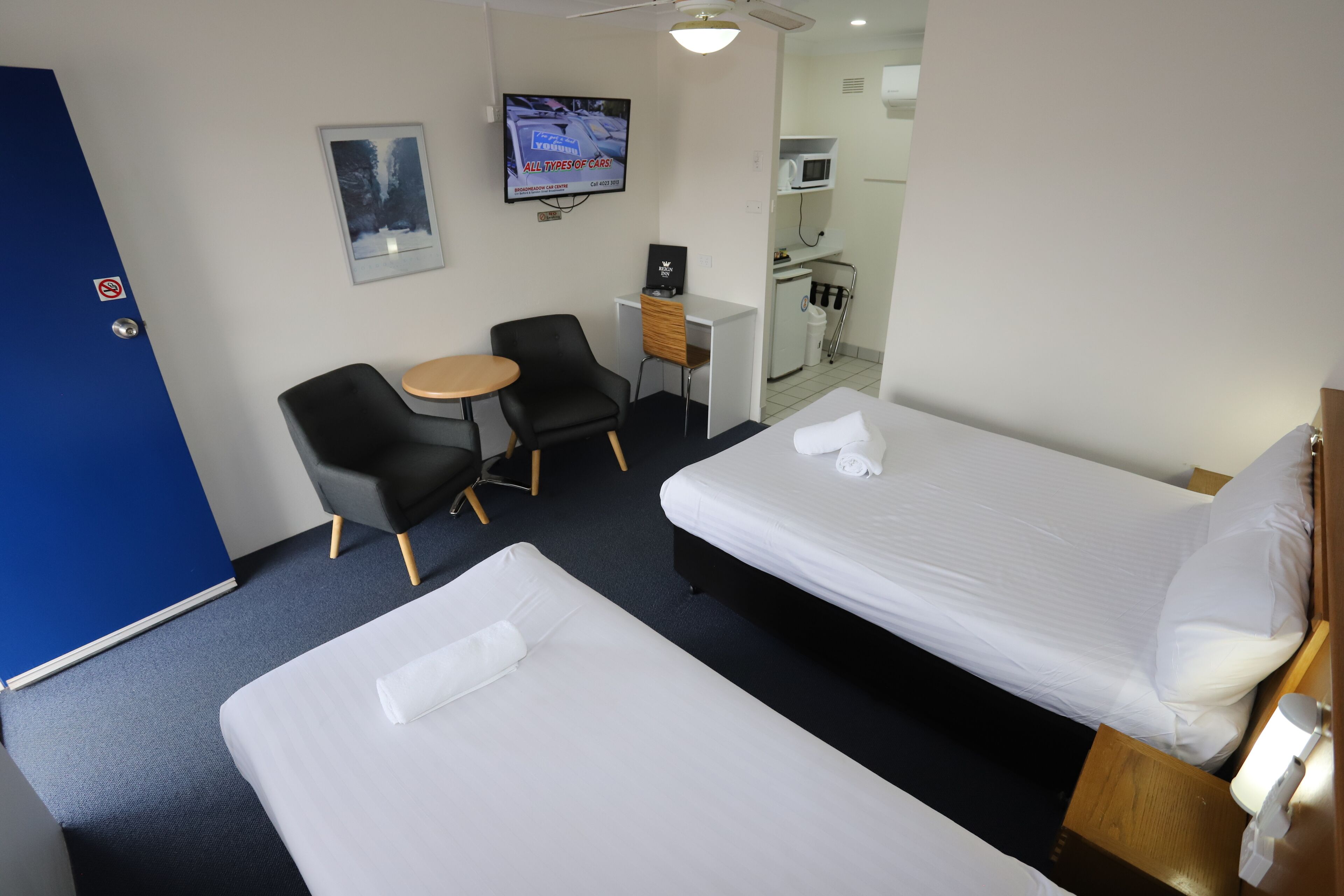 A hotel room with two beds, a desk, a chair, a TV, and a small kitchenette.