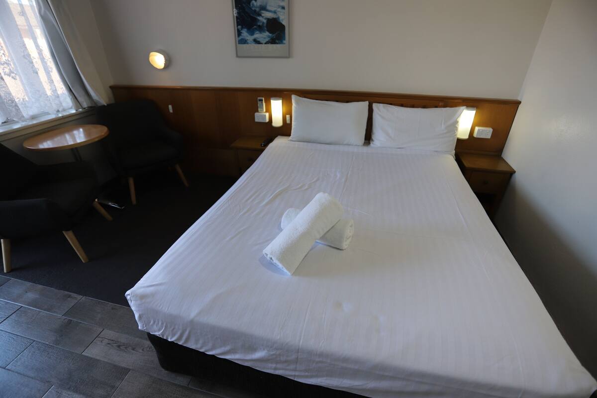A neatly made bed with white linens and a rolled towel, a small round table with a chair, and a wall-mounted light fixture.