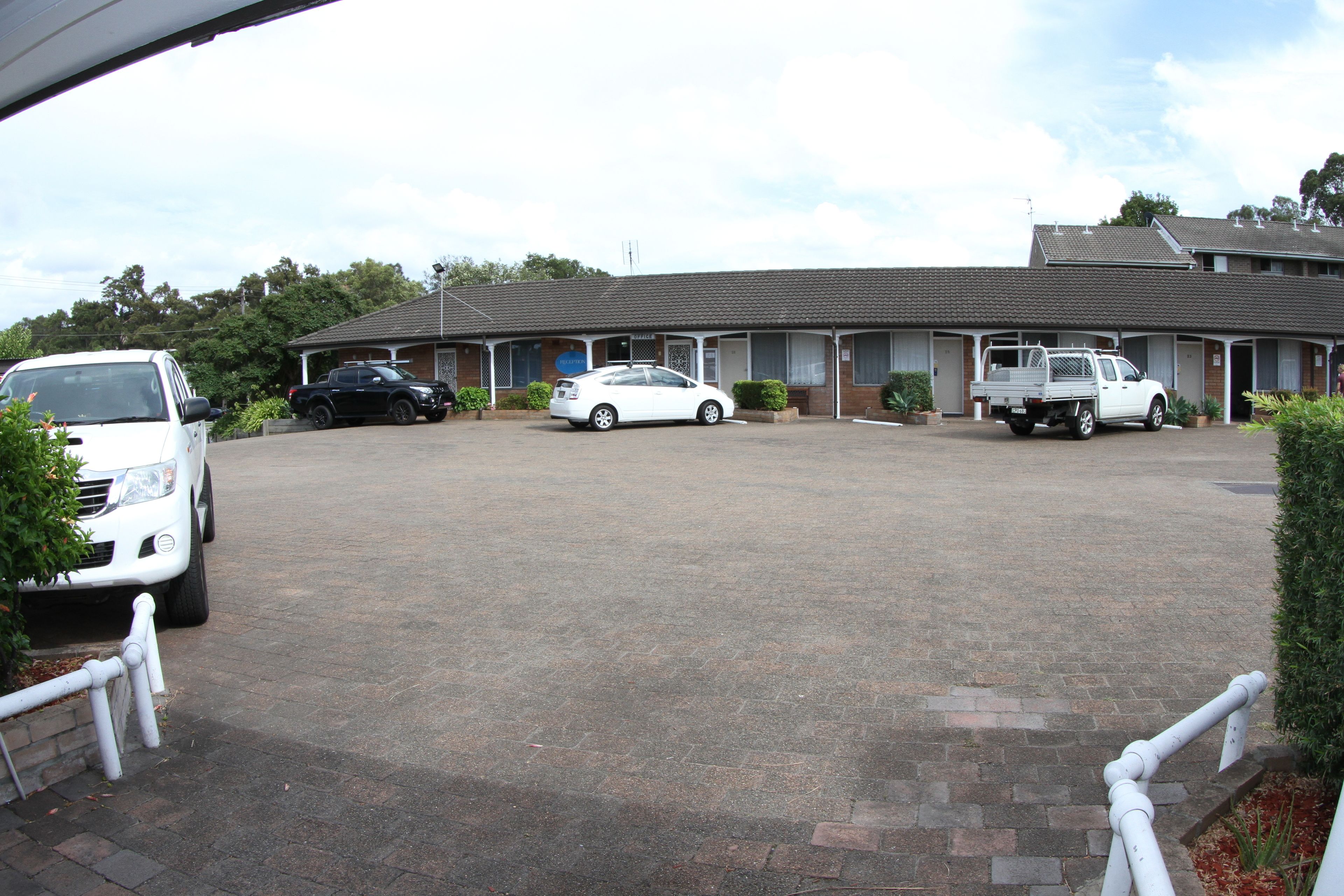 Un motel con un camino pavimentado, un automóvil blanco, un automóvil negro y una camioneta blanca estacionados al frente.