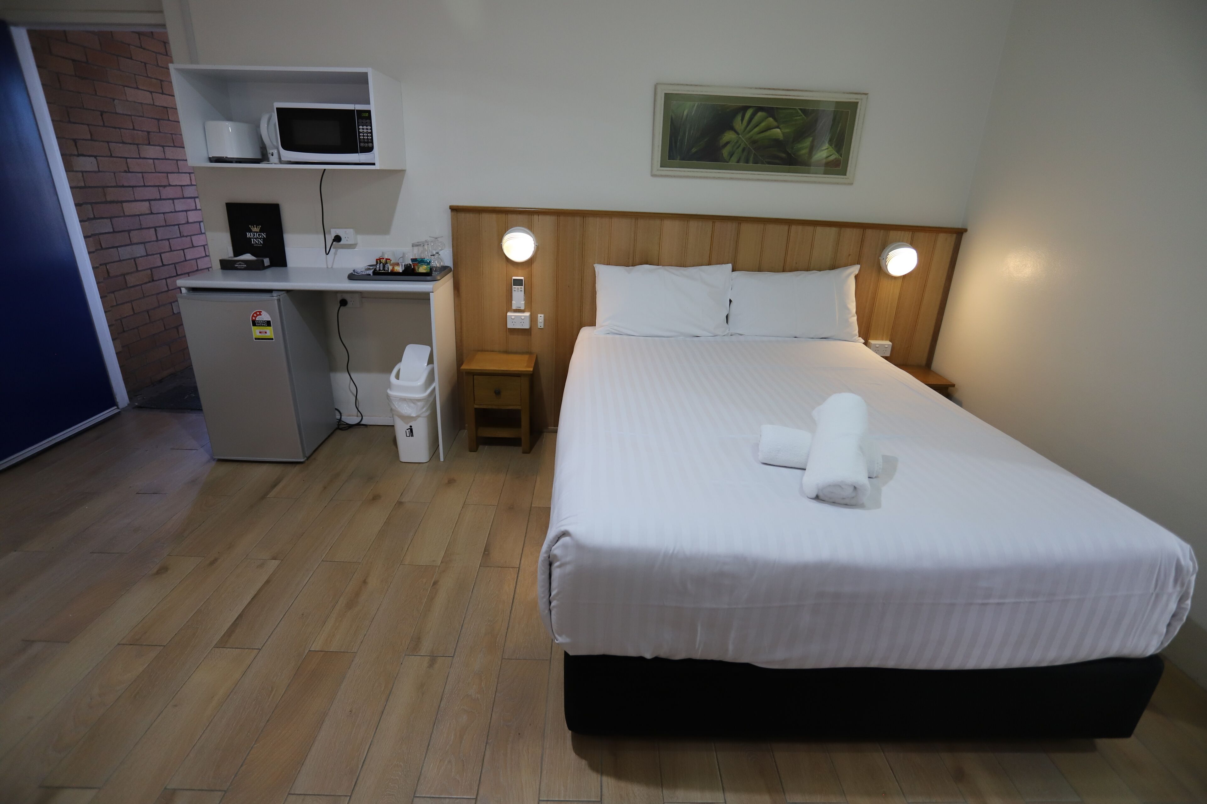 A hotel room with a bed, a desk with a microwave, a small refrigerator, and a nightstand with a lamp.