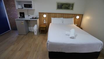 A hotel room with a bed, a desk with a microwave, a small refrigerator, and a nightstand with a lamp.