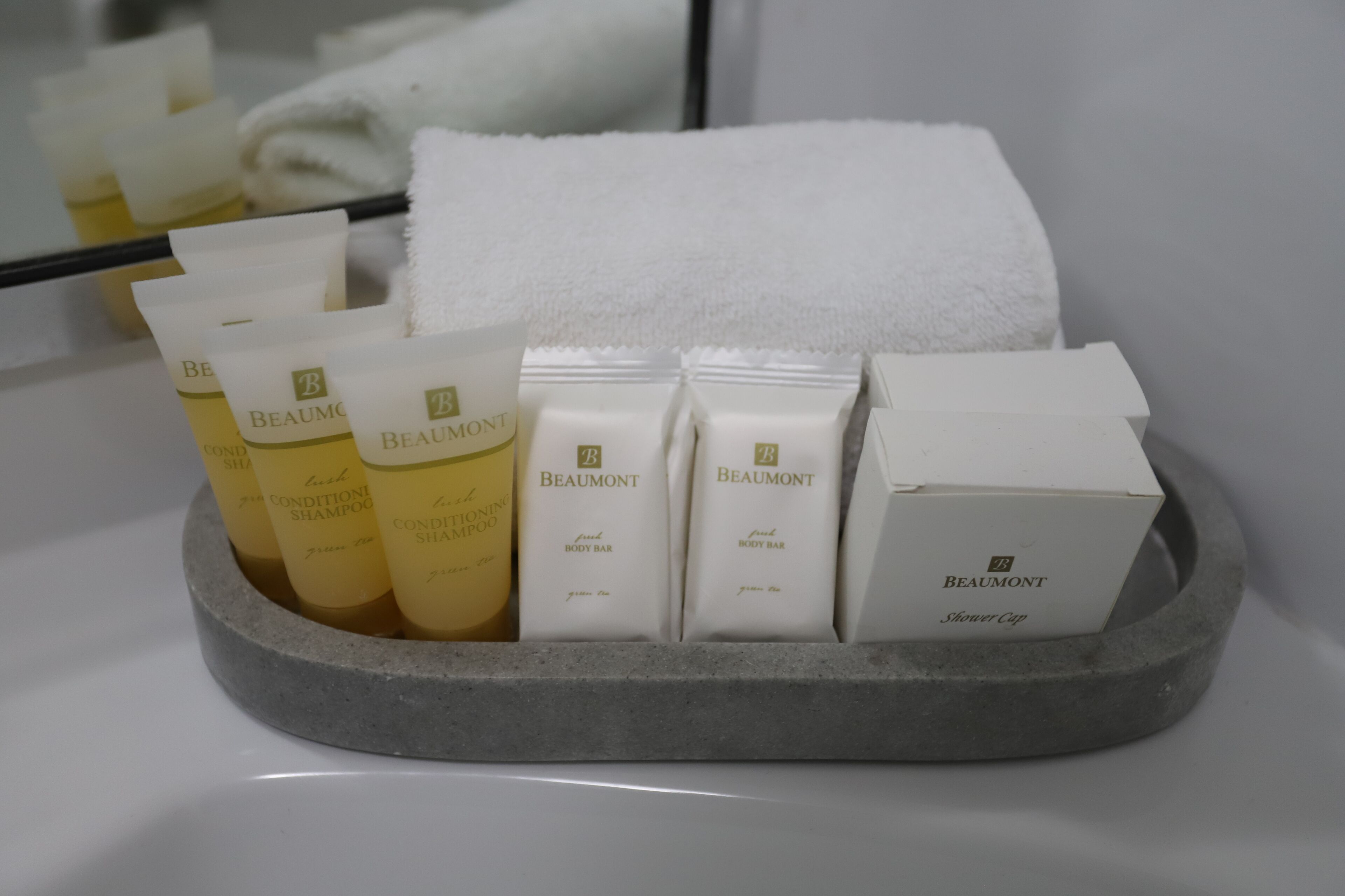 A bathroom with a grey stone tray holding various Beaumont toiletries including shampoo, body bar, and shower cap.