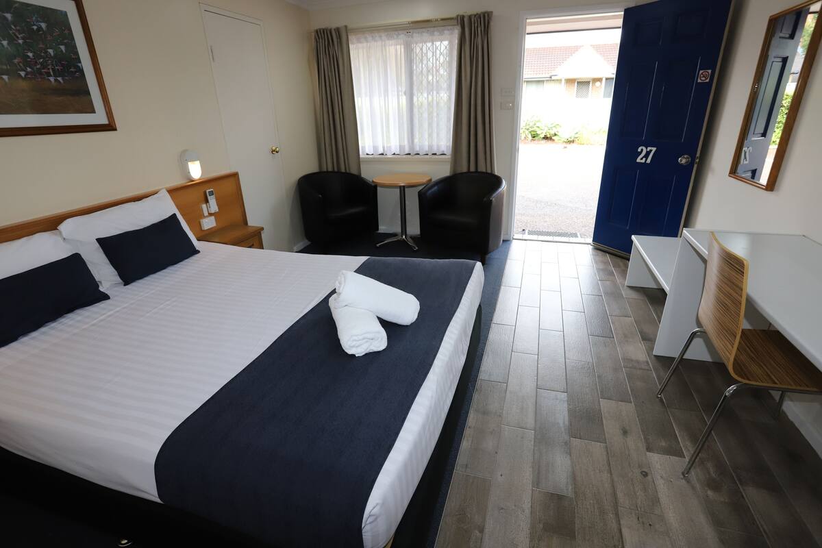 A hotel room with a bed, bedside table, lamp, two chairs, a desk, and a door.
