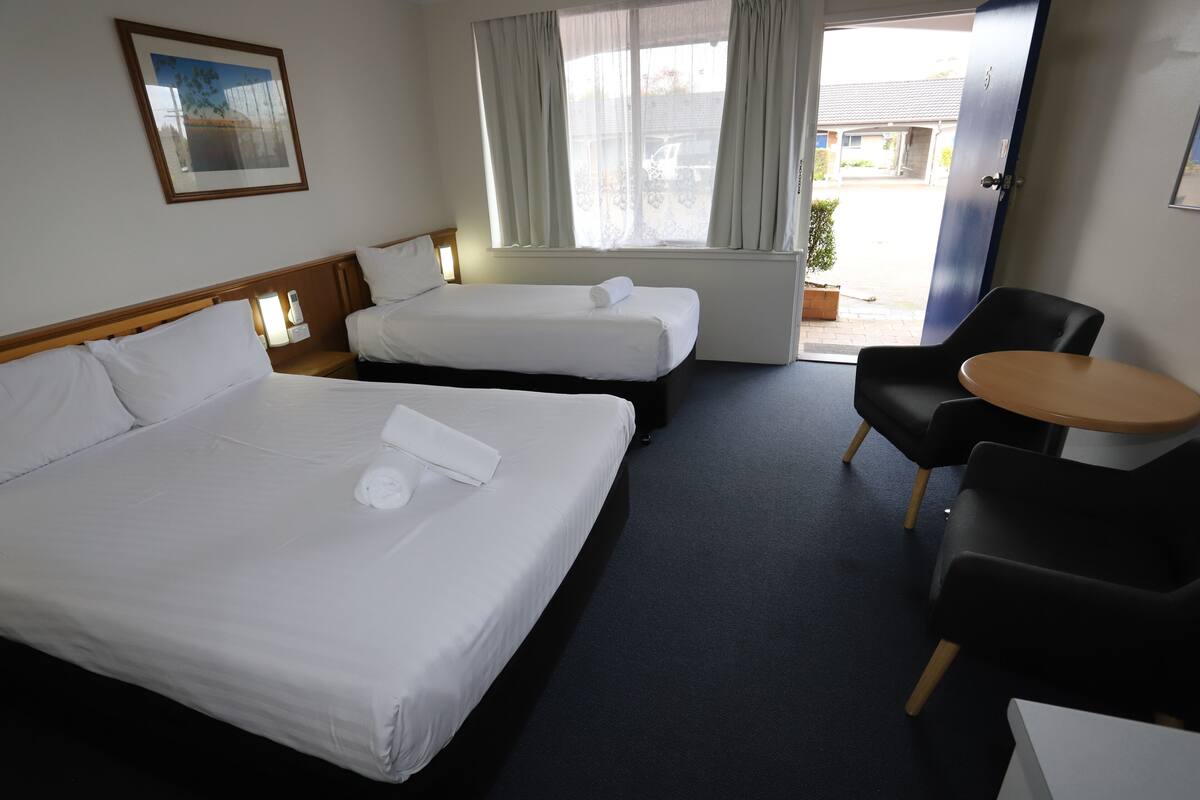 A hotel room with two beds, a small table, a chair, and a window with curtains.