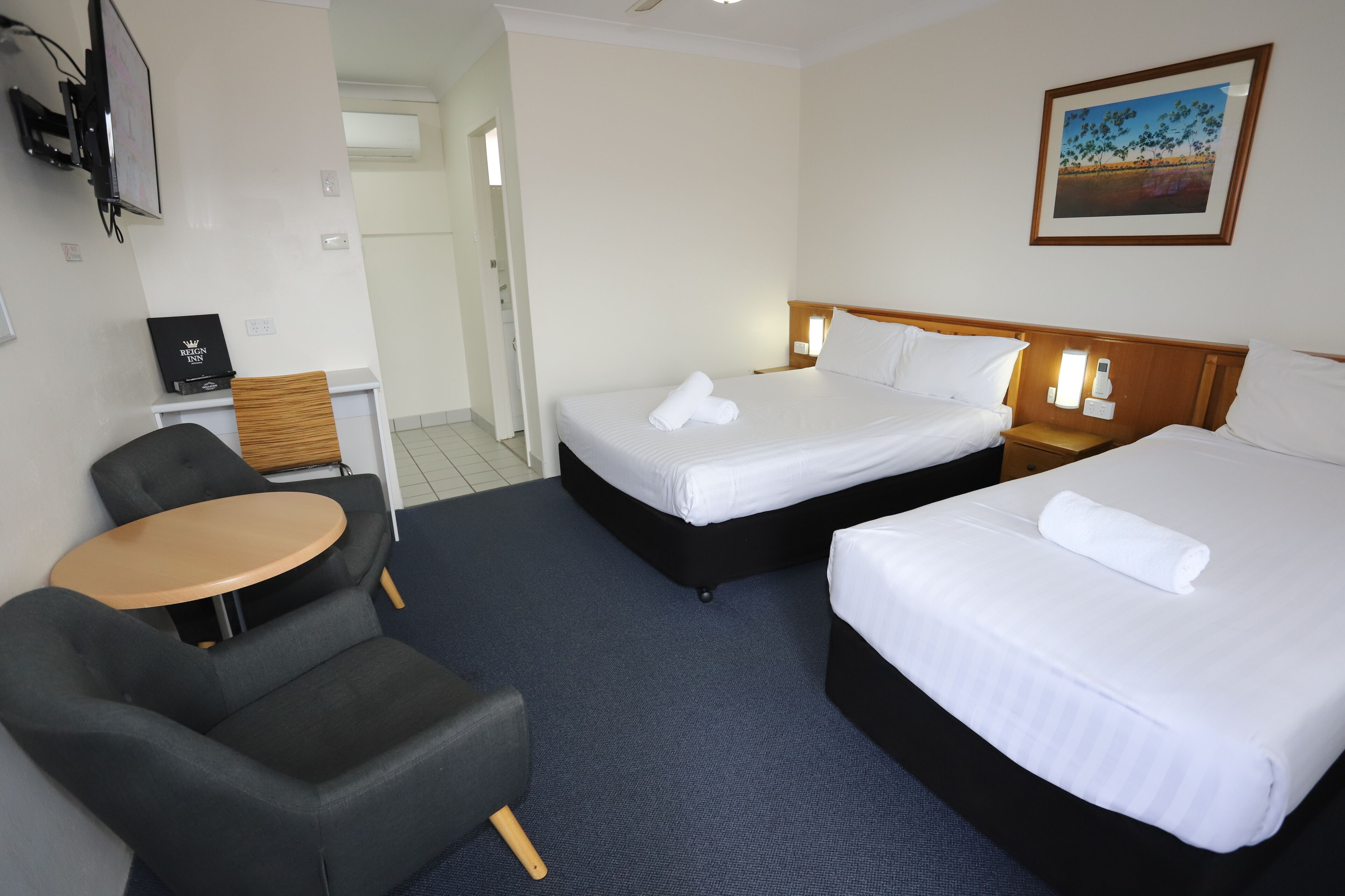 A hotel room with two beds, a small round table, a chair, and a painting on the wall.