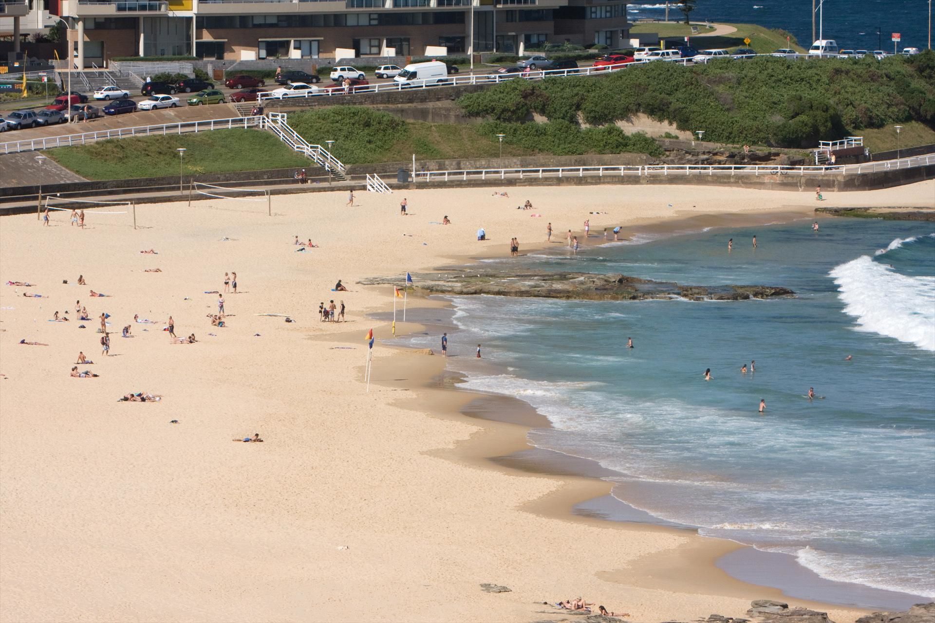 Une plage où des gens se prélassent au soleil et se baignent, un parking avec des voitures, et un bâtiment en arrière-plan.