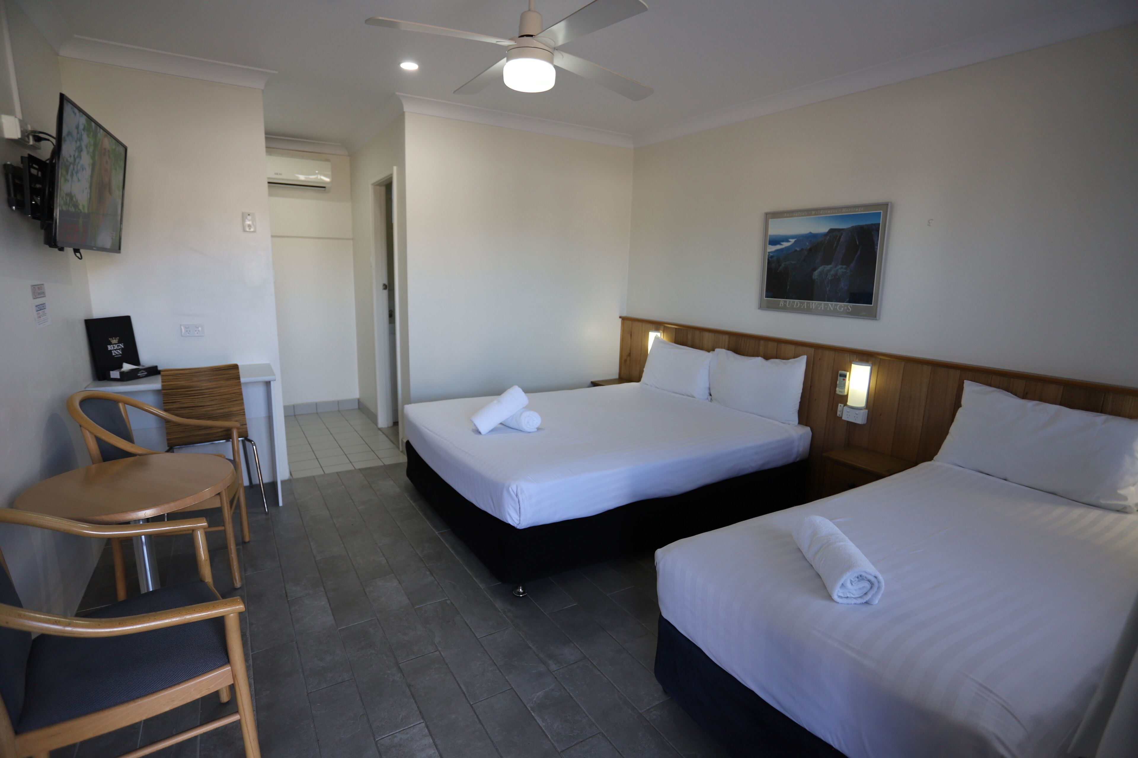 Habitación de hotel con dos camas, un escritorio con silla, un ventilador de techo y un cuadro en la pared.