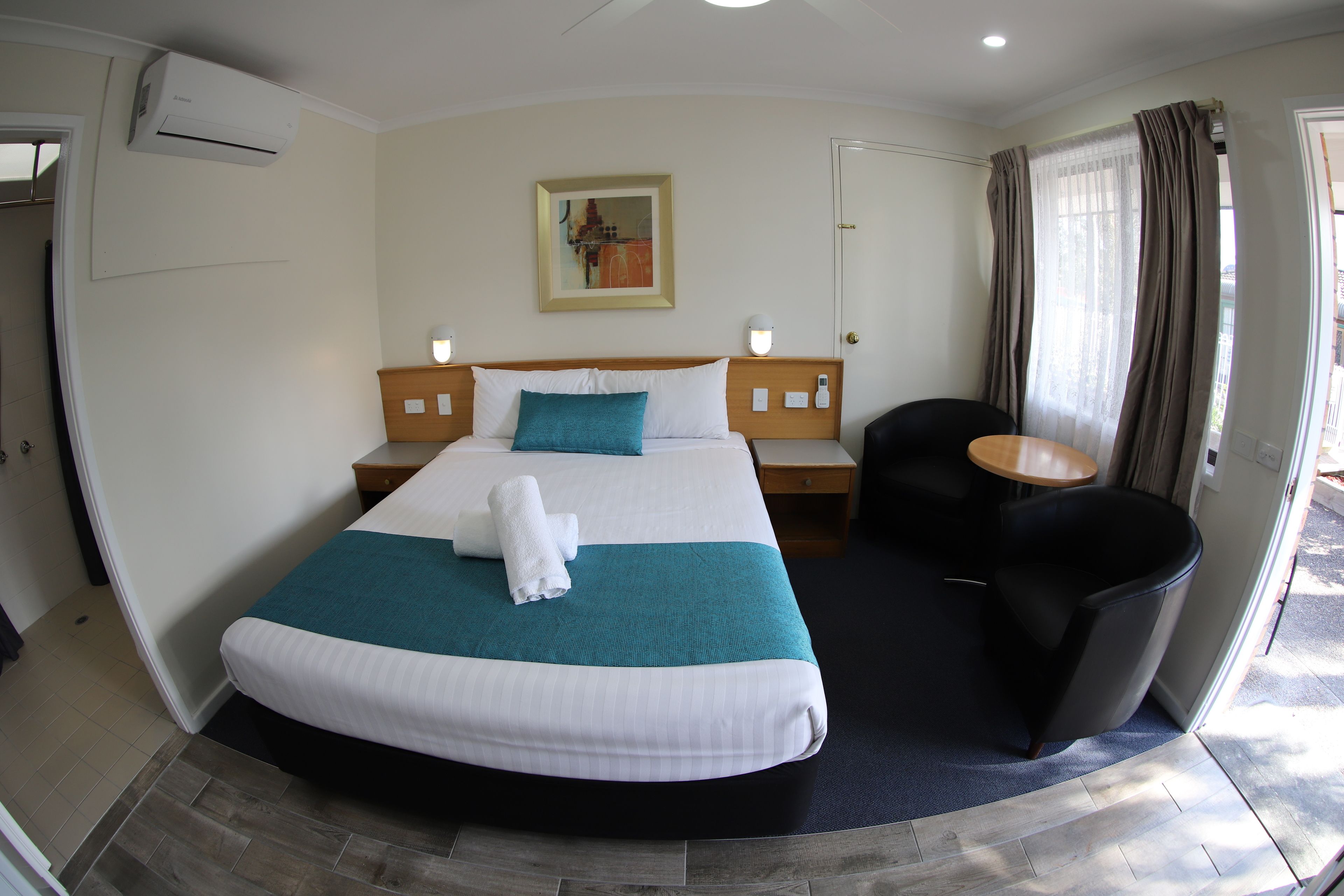 A hotel room with a bed, a chair, a small table, and a painting on the wall.