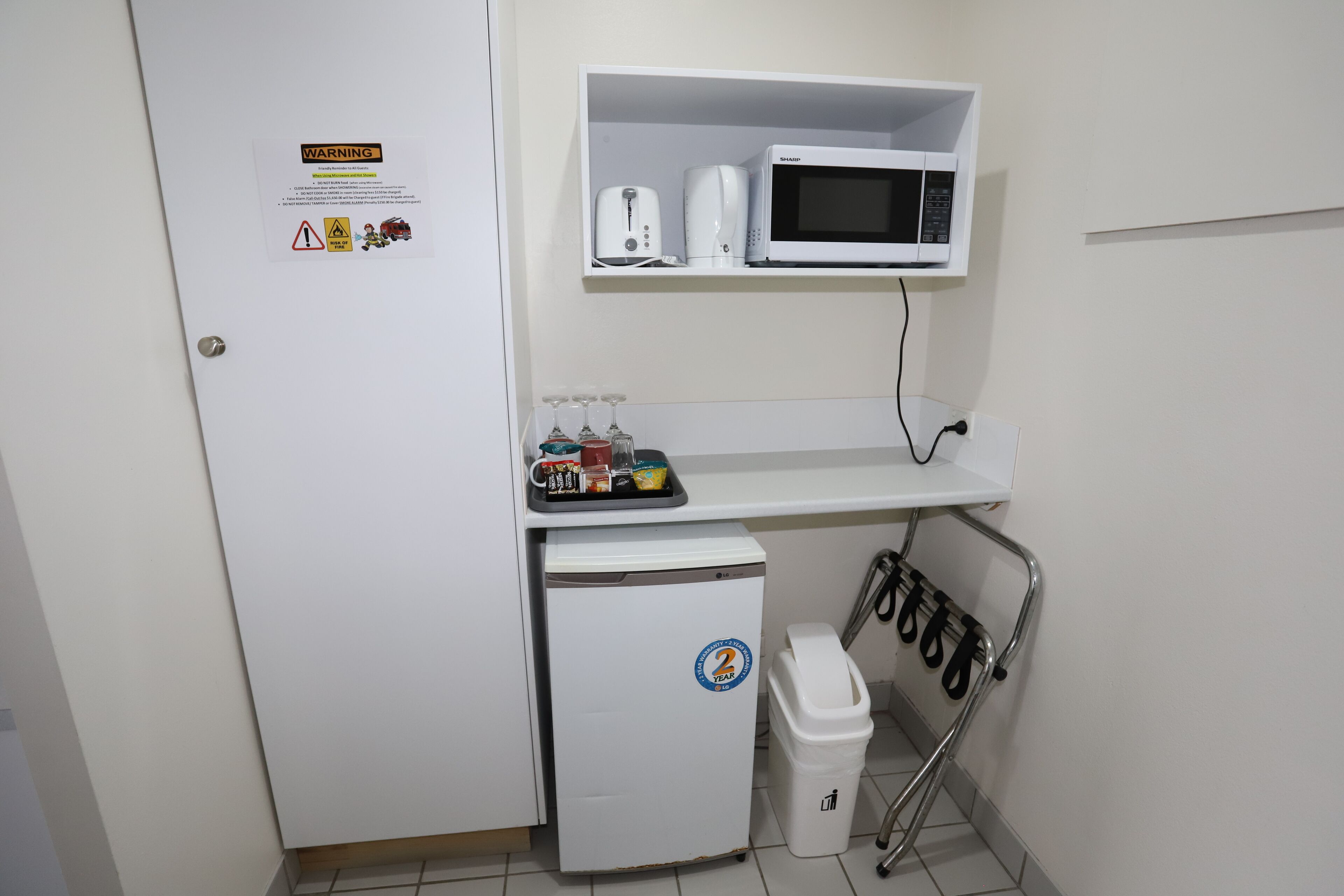 A compact kitchenette with a microwave, toaster, and small fridge. A trash bin and a shoe rack are also visible.