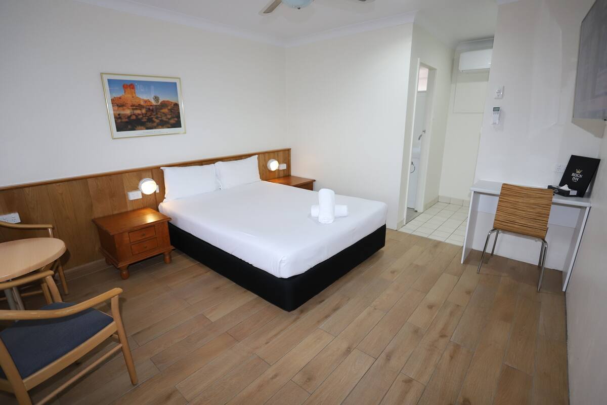 A hotel room with a bed, bedside tables, a desk, a chair, and a painting on the wall.