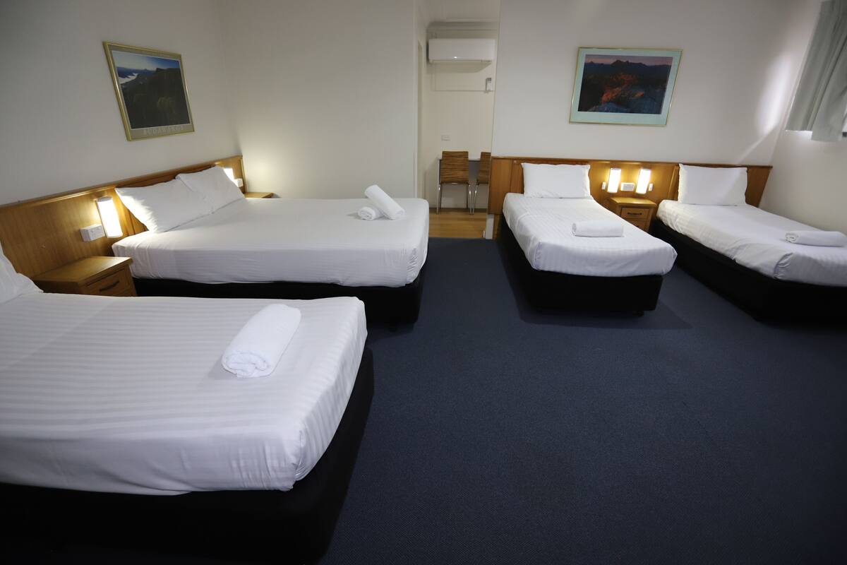 A hotel room with four single beds, each with a rolled towel, wooden headboards, and bedside tables with lamps.