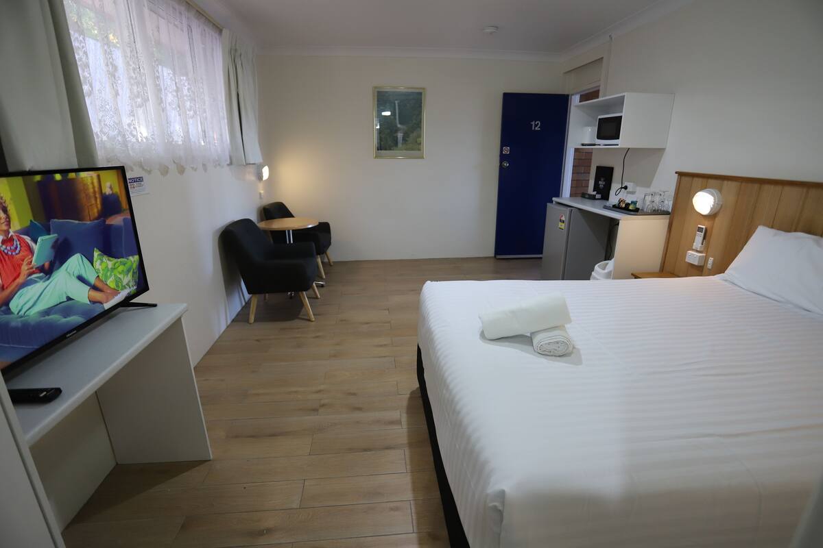A hotel room with a bed, a desk with a TV, a chair, a desk lamp, a microwave, and a door with a number.