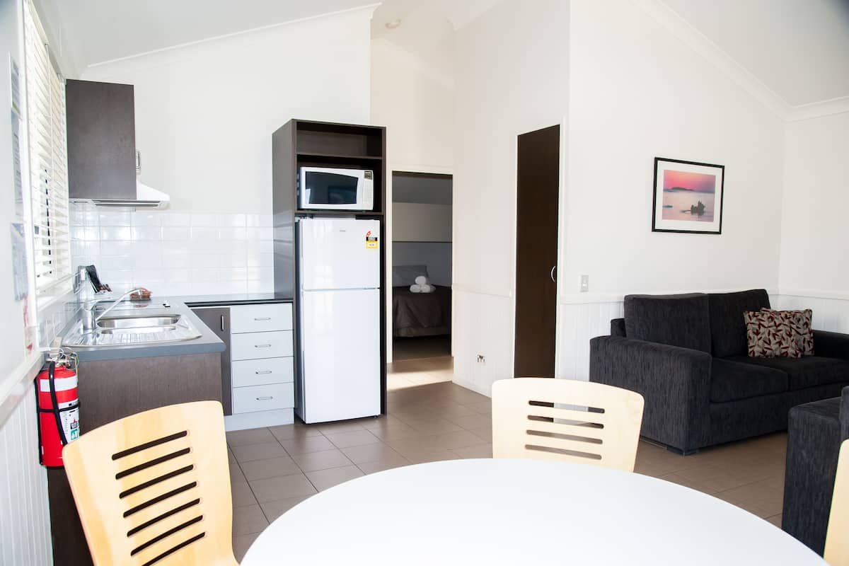 Una cocina moderna con microondas, refrigerador y lavaplatos. Hay un comedor con mesa y sillas, y un sofá gris con un cojín. Un cuadro enmarca una imagen en la pared.