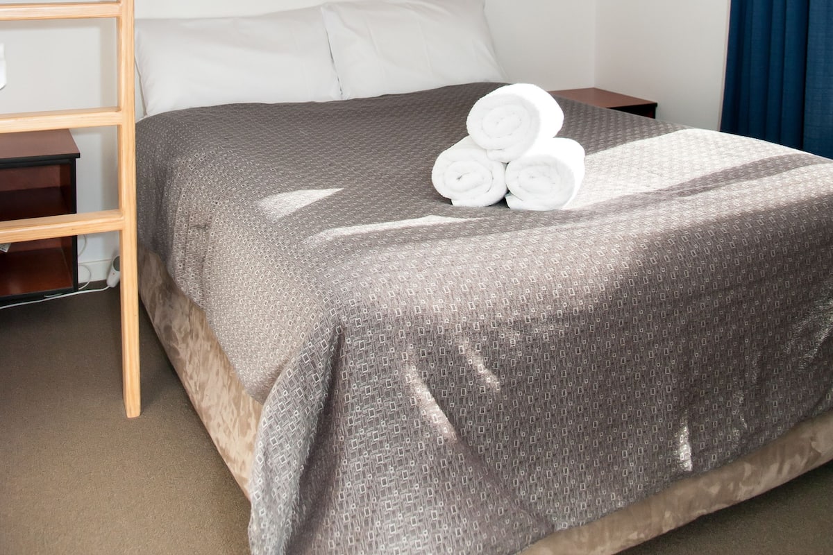 Una cama individual con un edredón gris estampado, almohadas blancas y toallas enrolladas sobre la cama. Una escalera de madera está a la izquierda de la cama, y una cortina azul se ve parcialmente a la derecha.