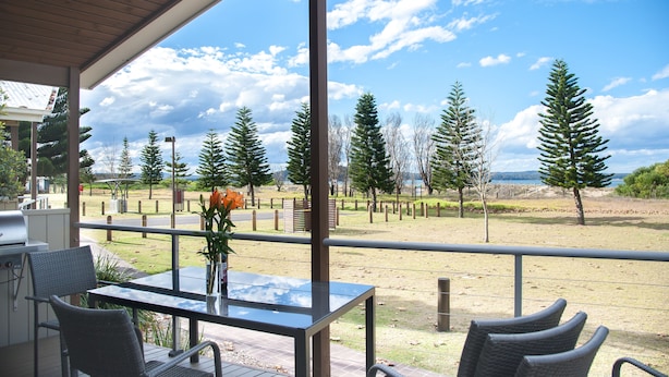 A covered outdoor area with a table and chairs, overlooking a grassy field with trees and a distant view of the ocean. - BIG4 Batemans Bay Beach Resort (Batehaven)