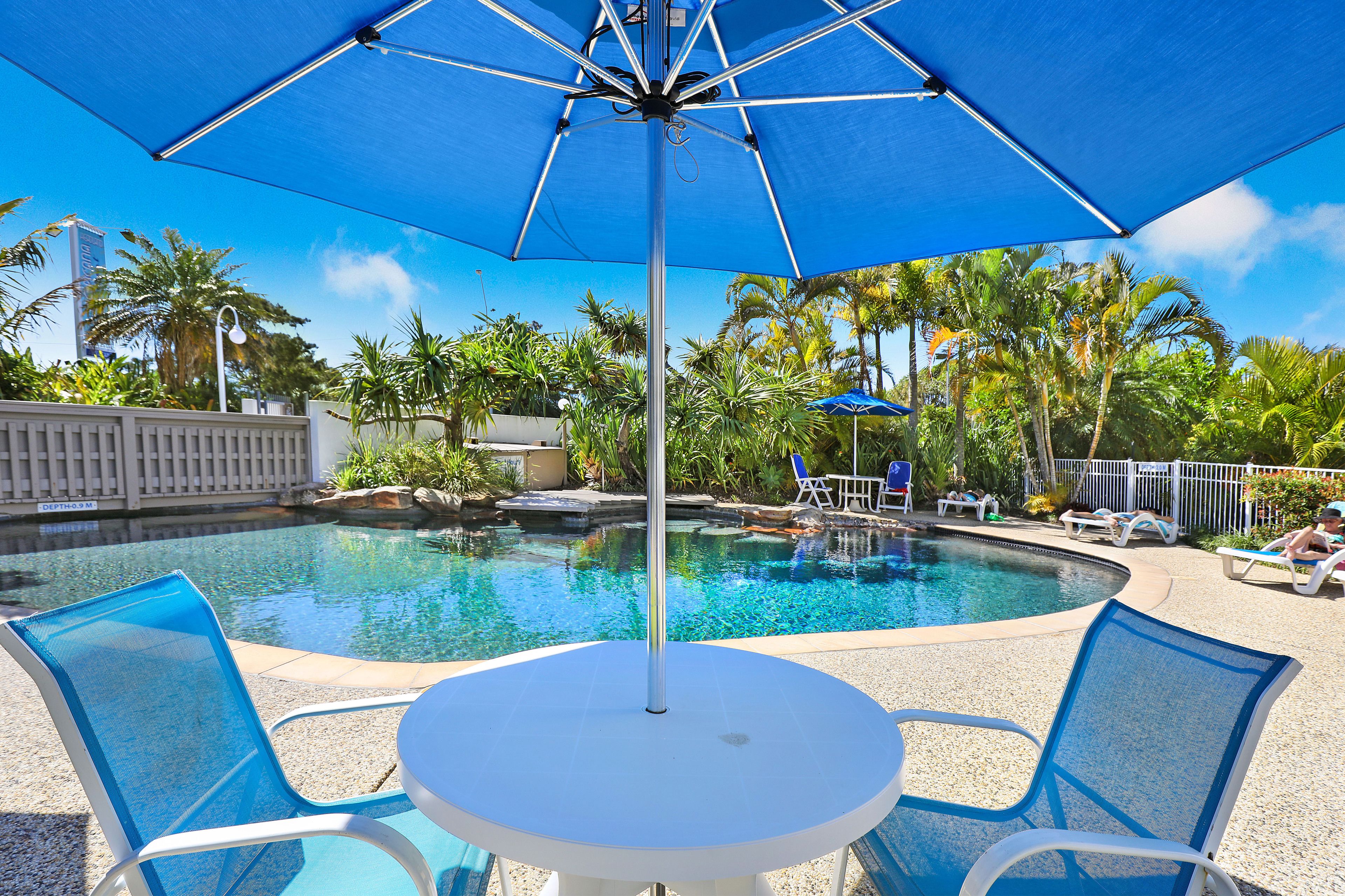 A pool area with a round table and chairs, a blue umbrella, and lounge chairs around a pool.