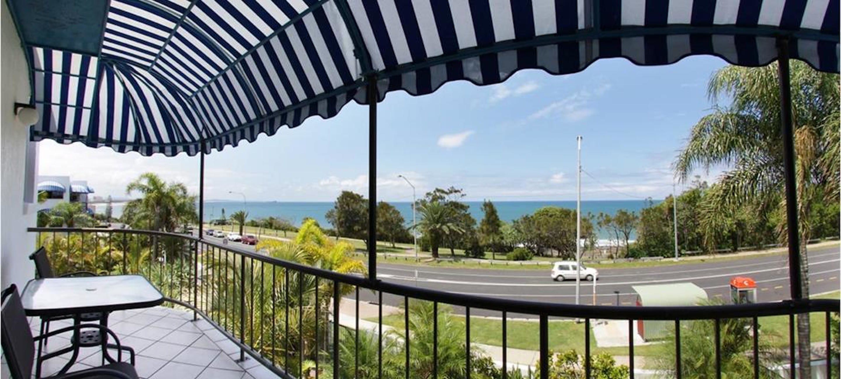 A balcony with a striped awning, a table, and chairs, overlooking a road and greenery with a distant view of the sea.