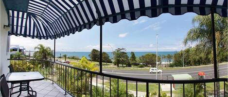 A balcony with a striped awning, a table, and chairs, overlooking a road and greenery with a distant view of the sea.