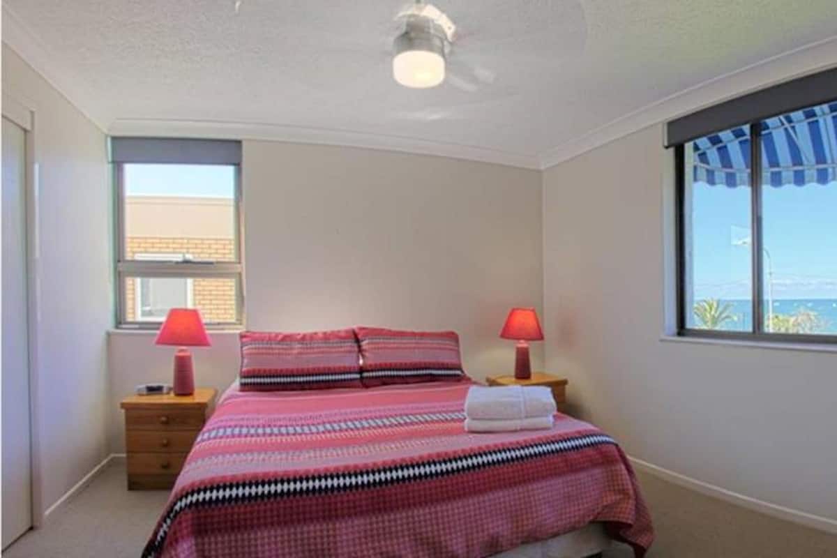 Un dormitorio con un ventanal grande, una cama con edredón a rayas, dos lámparas de noche y vista al mar.