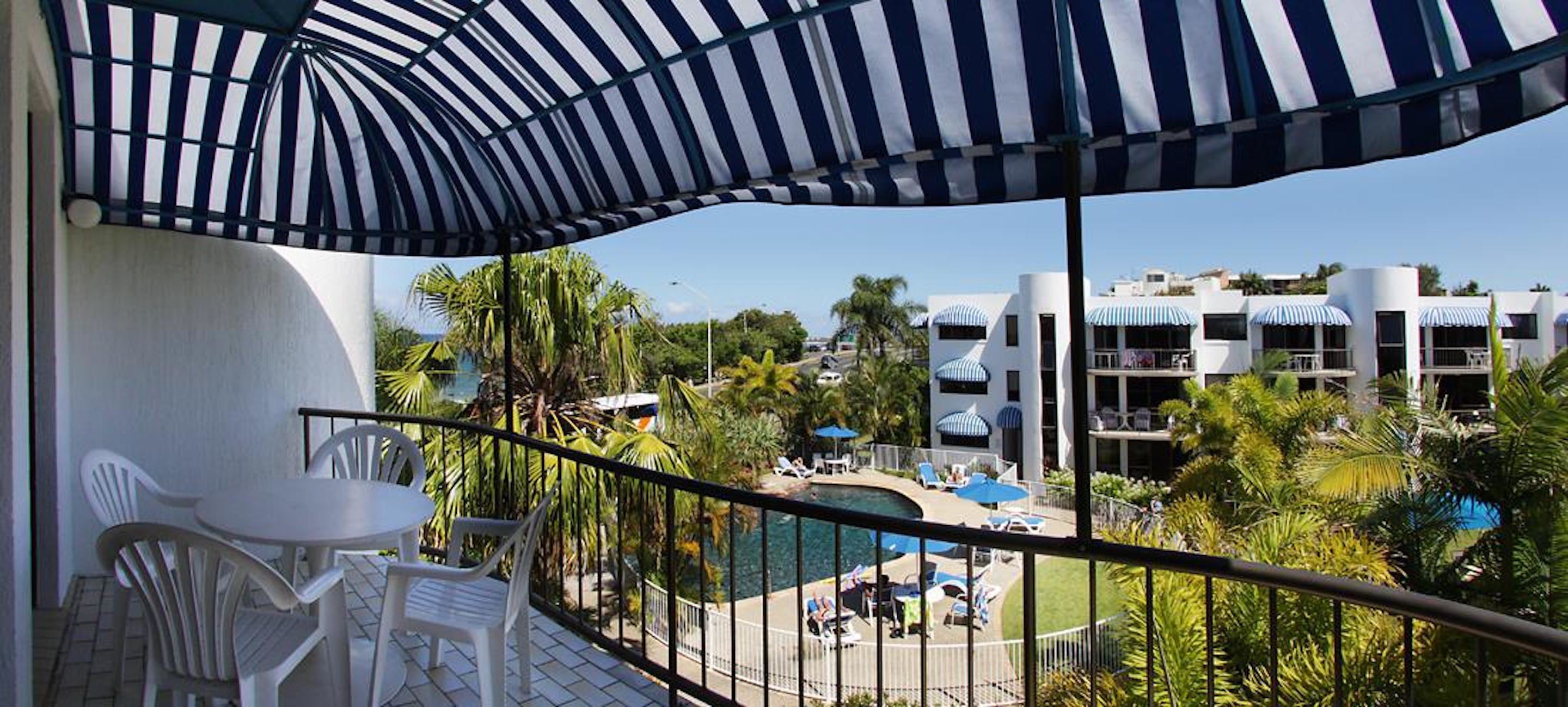 A balcony with a striped umbrella, white chairs, a pool area with lounge chairs, and a view of a resort with multiple buildings and palm trees.