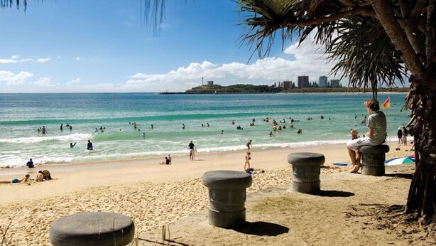 A beach with people swimming and sunbathing, palm trees, and a distant city skyline.