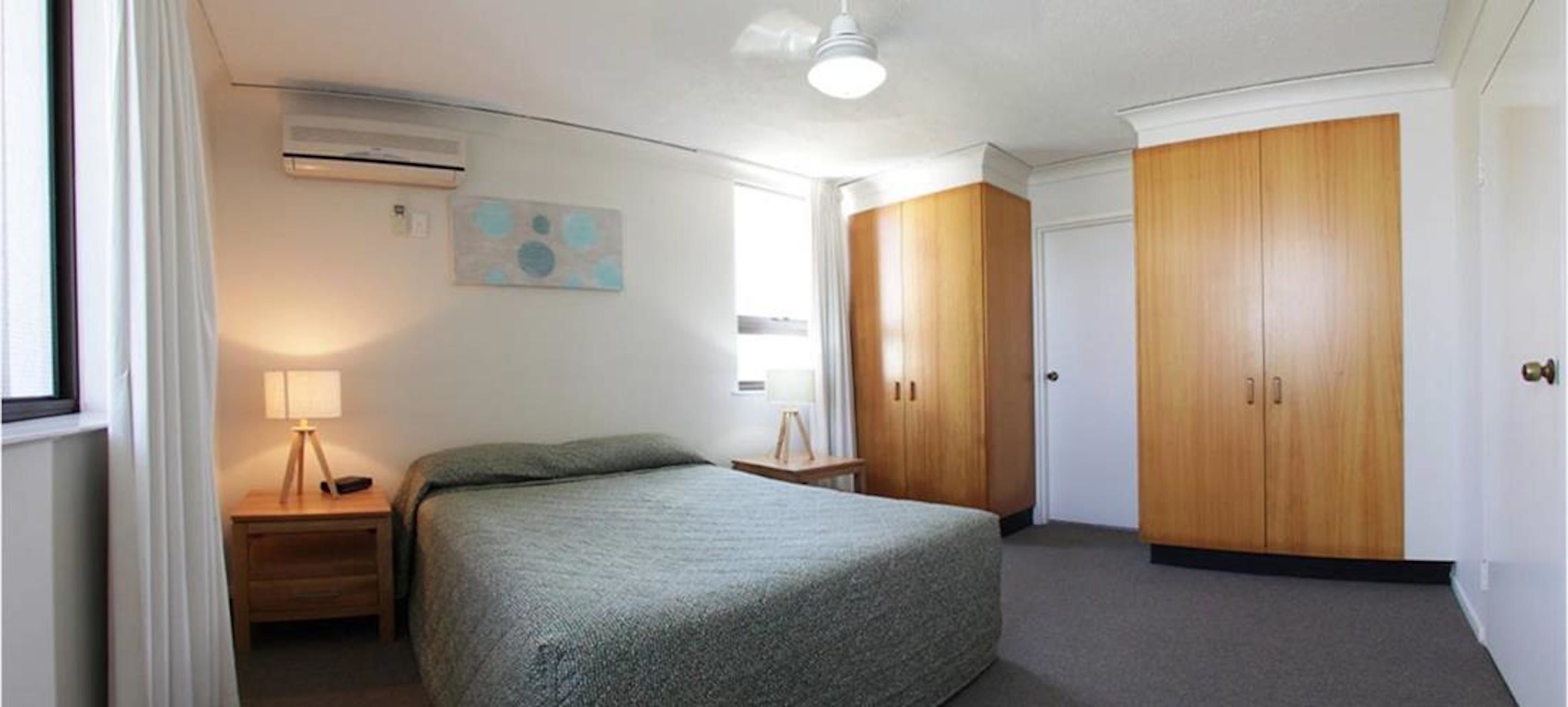Un dormitorio con cama, mesita de noche, unidad de aire acondicionado, armario de madera y un cuadro en la pared.
