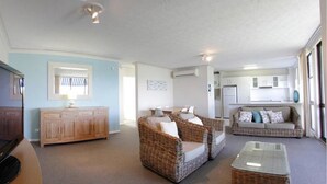 A living room with a flat-screen TV, wicker furniture, a wooden cabinet, and a kitchen area in the background.