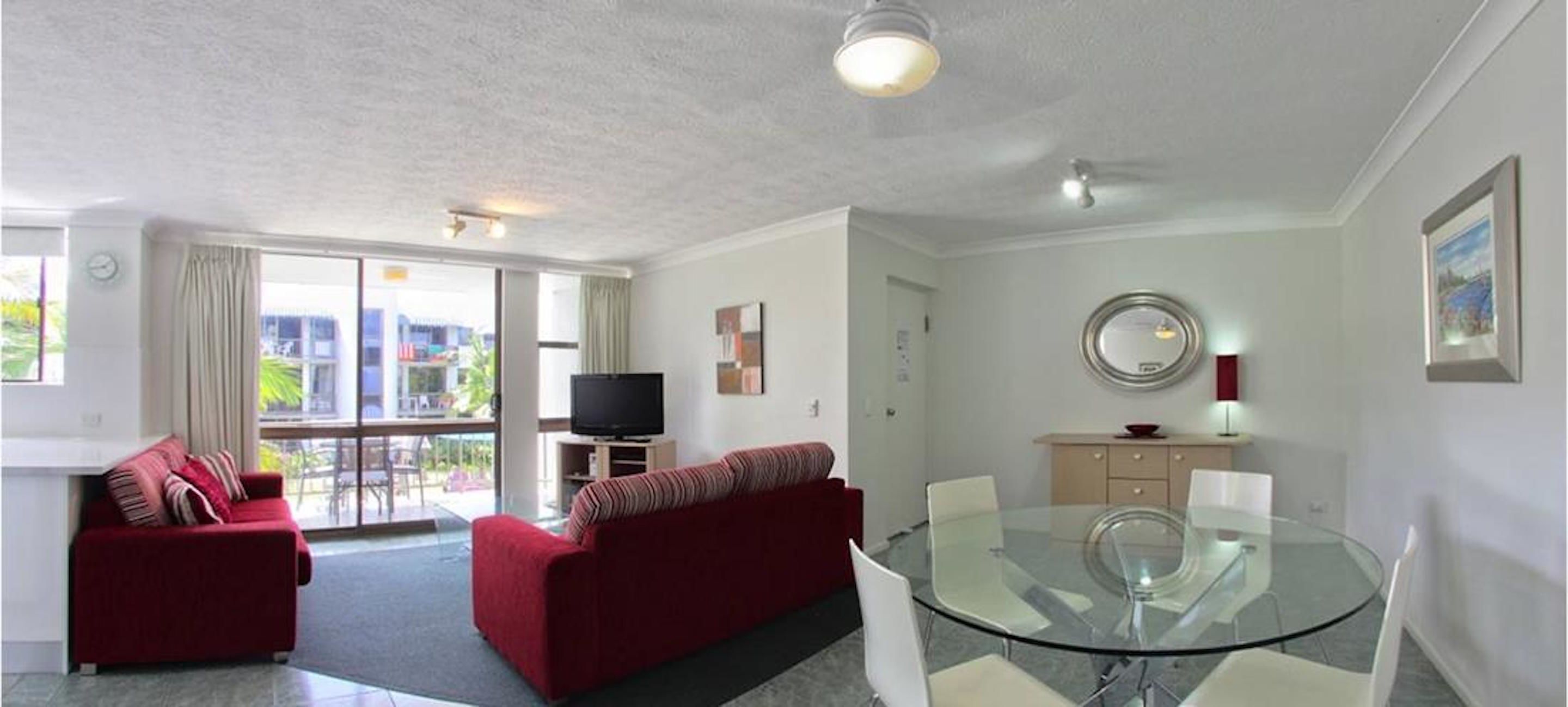 Una sala moderna con una mesa de comedor de cristal, sofás rojos, un televisor de pantalla plana y un balcón con vistas.