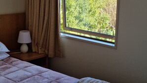A hotel room with a bed, a nightstand with a lamp, a window with a view of a mountain and greenery. - Methven Motels & Apartments (Methven)