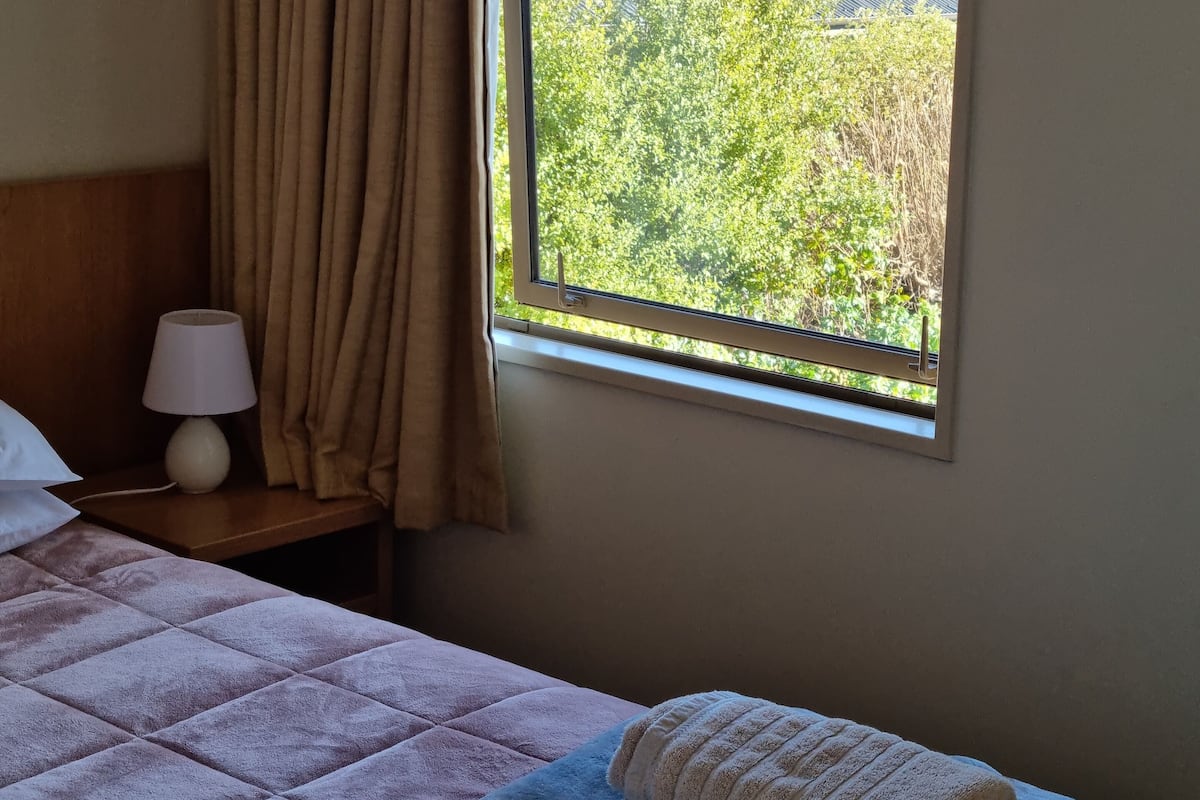 Habitación de hotel con cama, mesita de noche con lámpara, y ventana con vista a una montaña y vegetación.