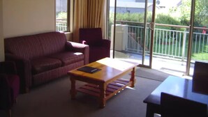 A room with a sofa, a chair, a table, and a view of greenery and buildings through large windows. - Methven Motels & Apartments (Methven)
