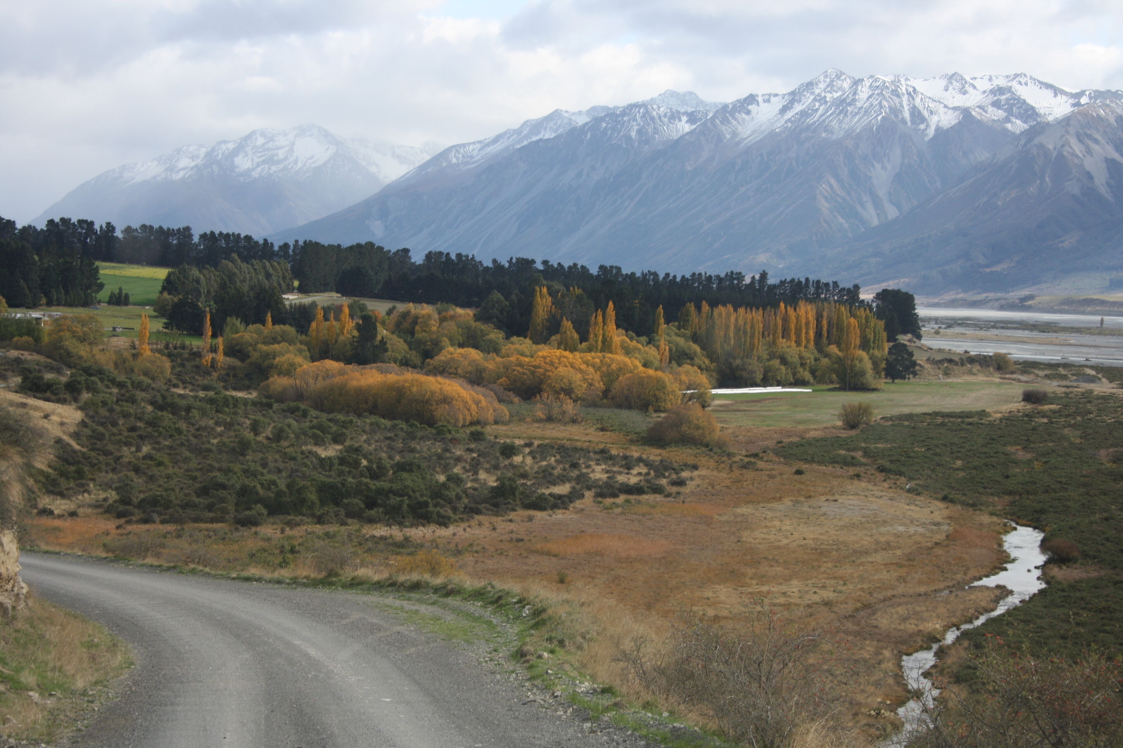 A rural landscape with a winding road, a small stream, and a forested area with autumn-colored trees.