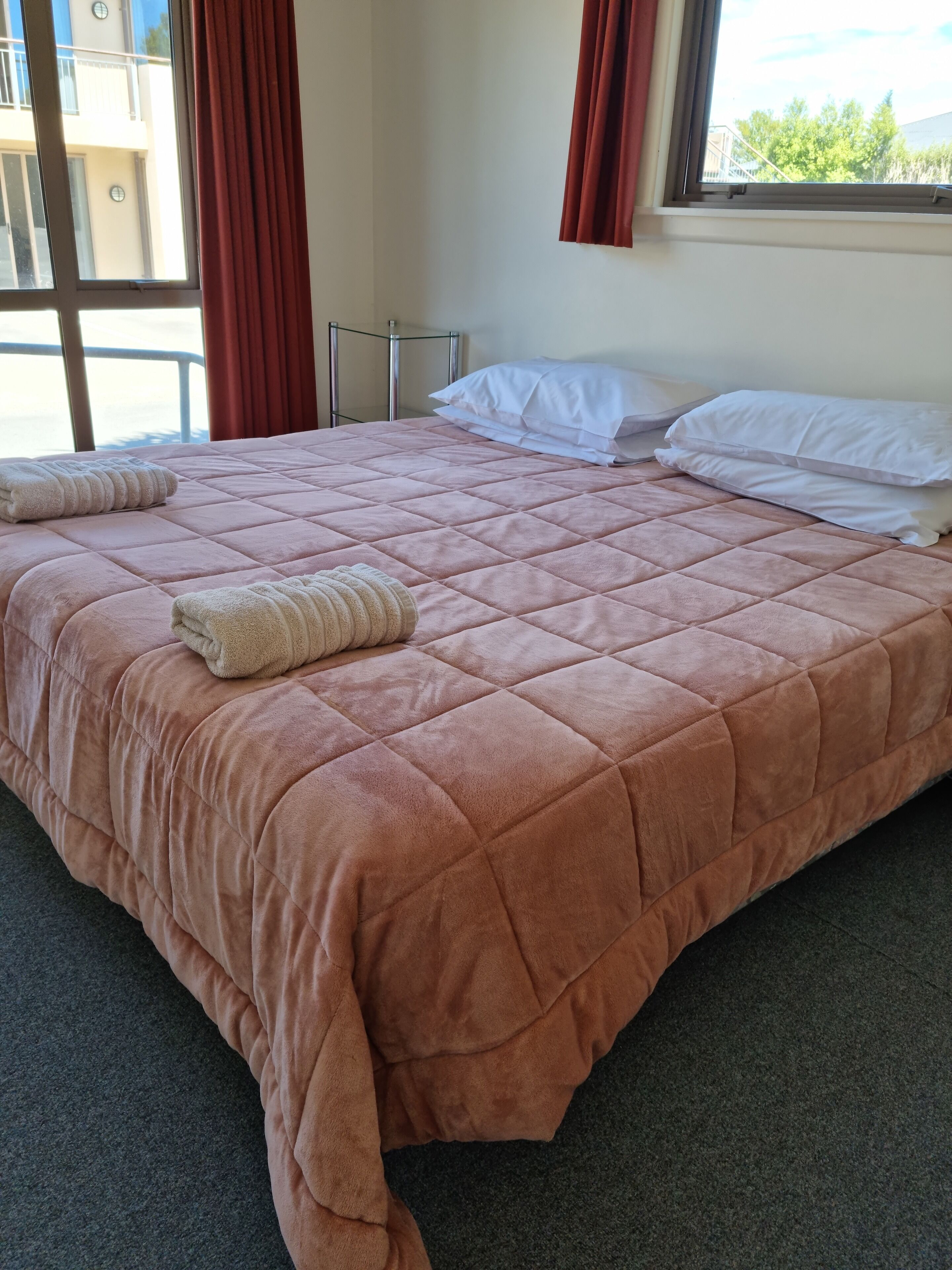Una cama bien hecha con colcha rosa, almohadas blancas y una toalla doblada.