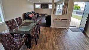 A spacious living area with a dark wood floor, a glass-top dining table with patterned chairs, a black leather sofa with red cushions, and a flat-screen TV mounted on the wall.