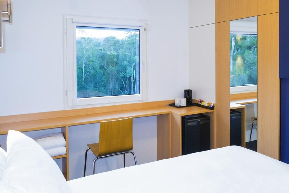 Une chambre d’hôtel équipée d’un lit, d’un bureau, d’une chaise, d’un petit réfrigérateur et d’une fenêtre donnant sur des arbres.