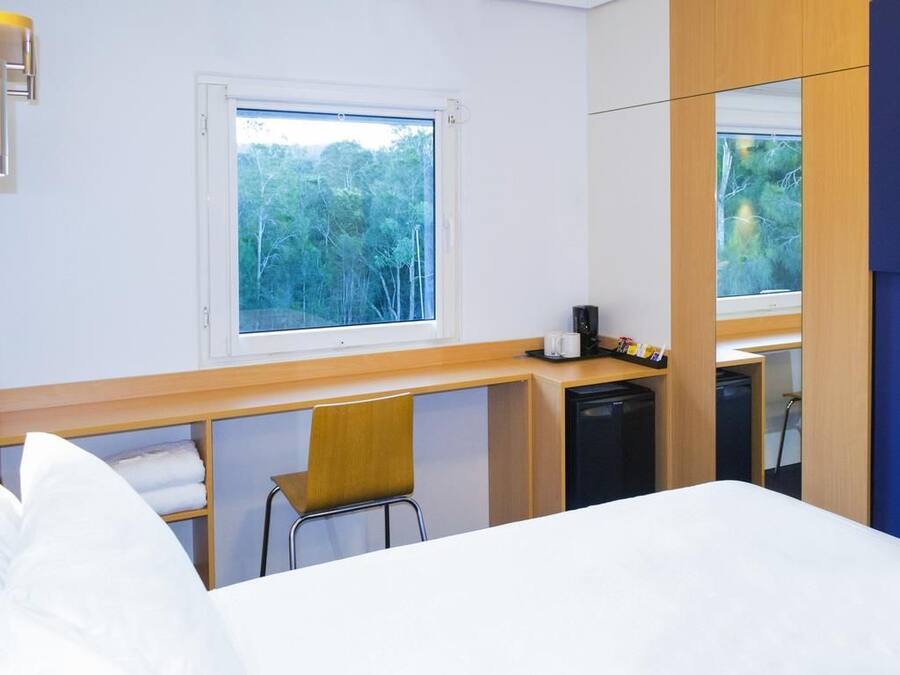 Ein Hotelzimmer mit Bett, Schreibtisch, Stuhl, kleinem Kühlschrank und einem Fenster mit Blick auf Bäume.