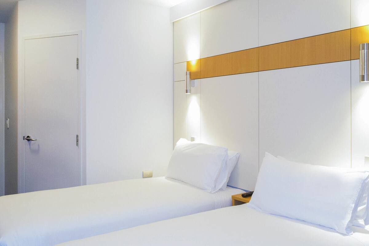 A hotel room with two single beds, a wooden panel on the wall, a wall-mounted light, and a door.