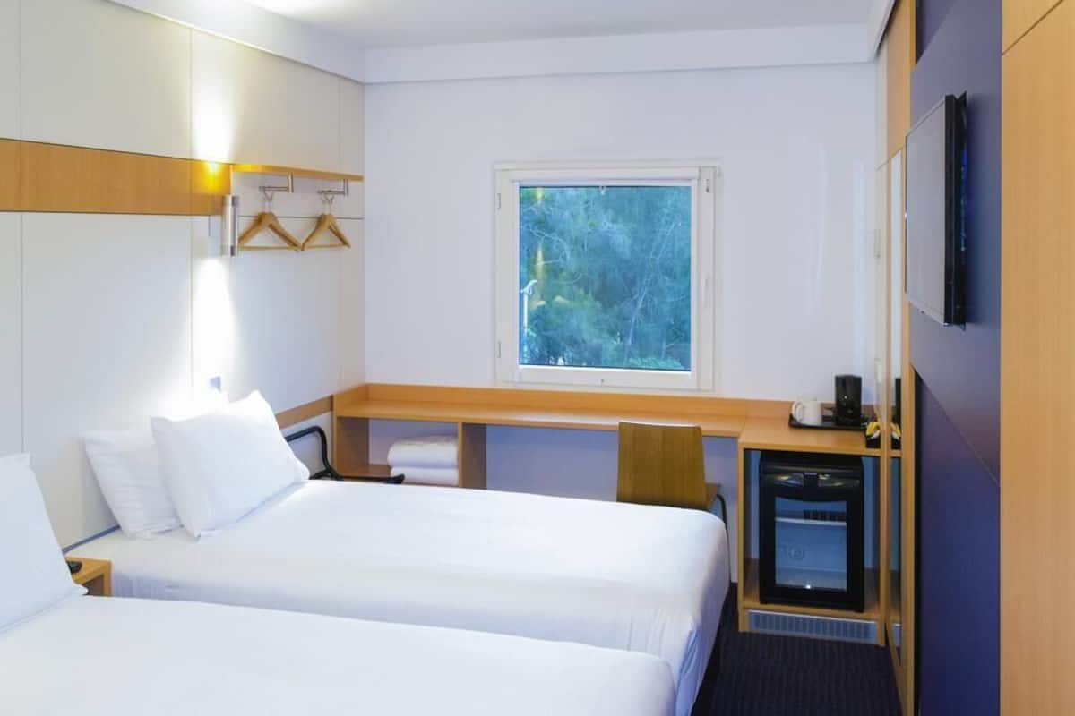 A hotel room with two beds, a desk, a TV, and a window with a view of trees.