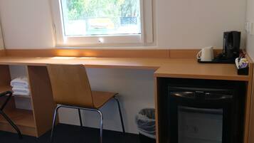 Habitación de hotel con un escritorio de madera, una silla, una máquina de café, un cesto de basura y una ventana con vistas a árboles.