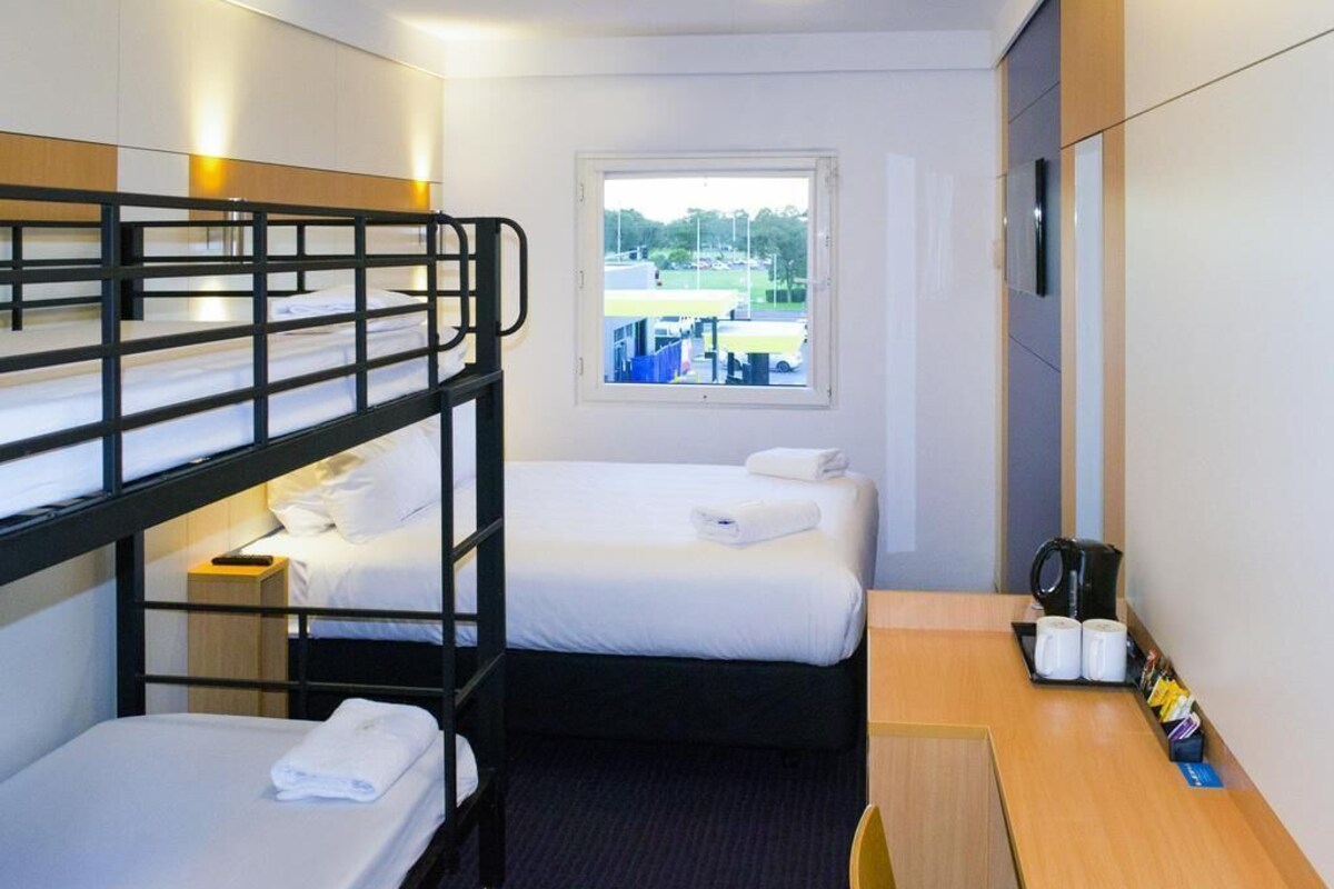 Hotelkamer met stapelbedden, een enkele bed, een bureau met waterkoker en een raam met uitzicht op een parkeerplaats.