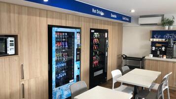 Um café moderno com máquinas de venda automática para bebidas quentes, lanches e bebidas frias, e uma área de estar com mesas e cadeiras.